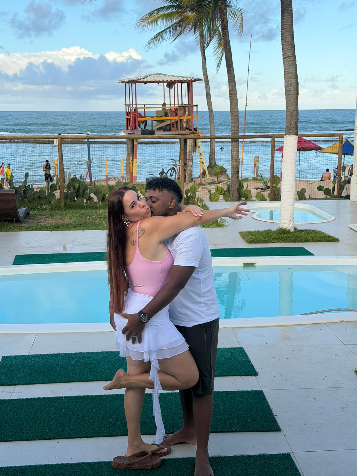 Greice e Ely abraçados à beira da piscina, com o mar ao fundo